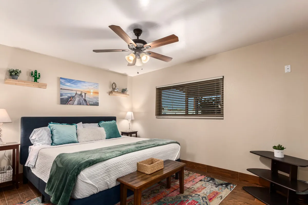 A cozy bedroom with a neatly made bed adorned with decorative pillows, artwork on the wall, a window with blinds letting in natural light, and shelves with potted plants. The room radiates comfort and relaxation, inviting restful sleep or lazy mornings.
