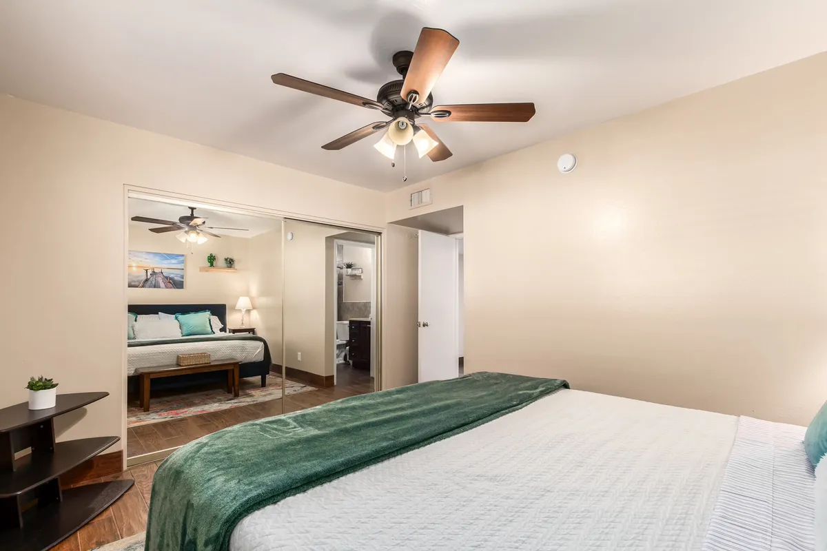 A serene bedroom featuring a bed with a green throw, a ceiling fan for comfort, decorative wall art, and a nightstand with a lamp. The space is bright yet tranquil, ideal for relaxation and a good night's sleep.