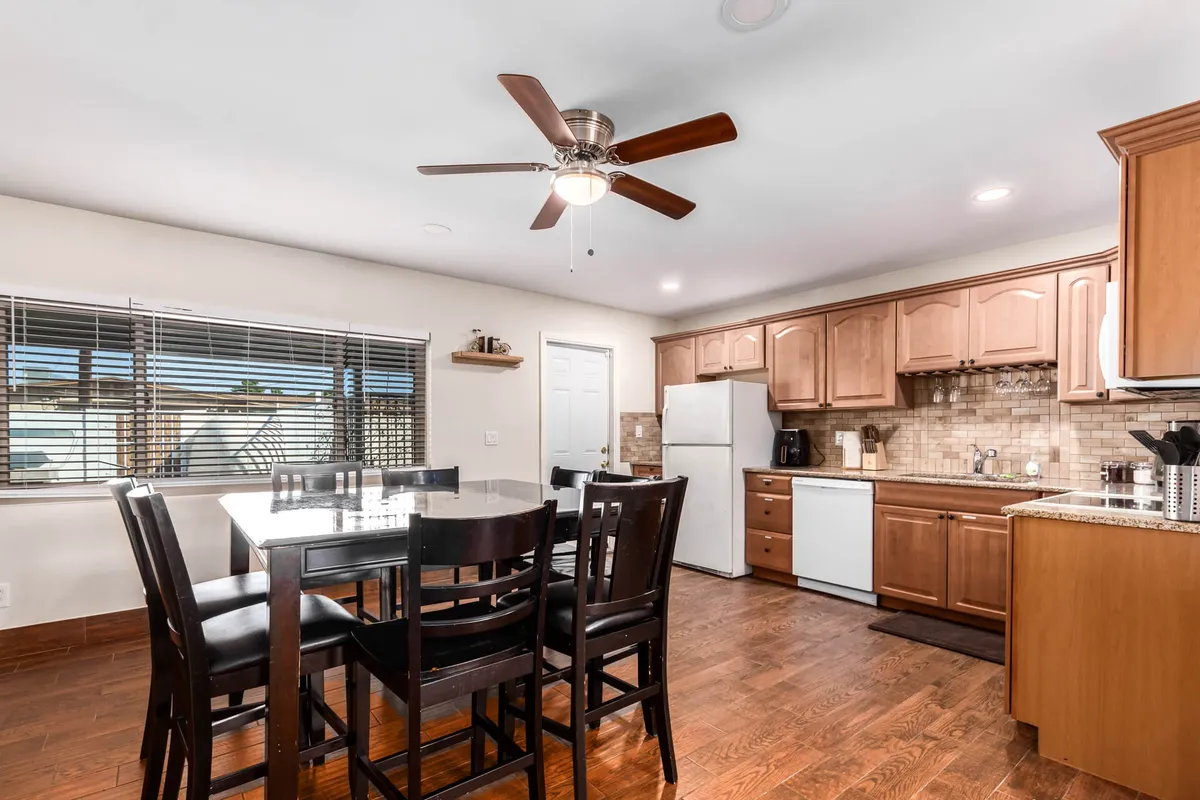 A dining table sits near the kitchen cabinets, with a large window letting in natural light and a ceiling fan overhead. The setup is ideal for family meals, hosting guests, or enjoying morning coffee in a warm and welcoming environment.