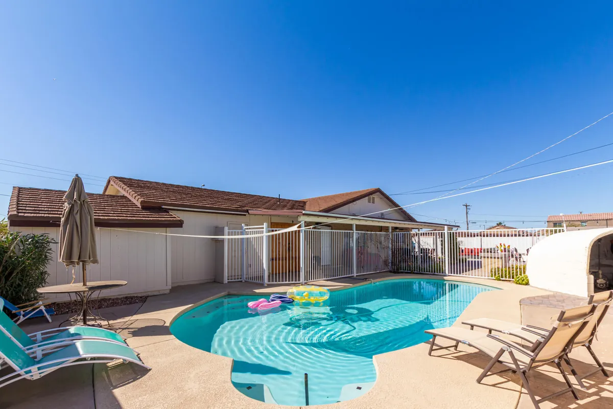 This image captures a bright, inviting backyard scene featuring a sparkling swimming pool surrounded by lounge chairs, an umbrella, and playful inflatable pool toys. The atmosphere is lively, with clear indications of children and families enjoying the water and sunshine. The setting is perfect for socializing and relaxation.