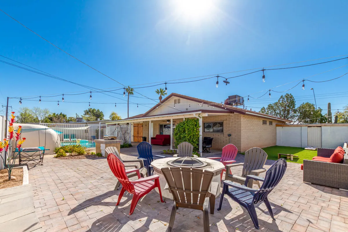 This image centers on a cozy fire pit area with outdoor seating, the pool in the background, and string lights overhead. The space is set for barbecuing, pool parties, and relaxed evening gatherings around the fire.