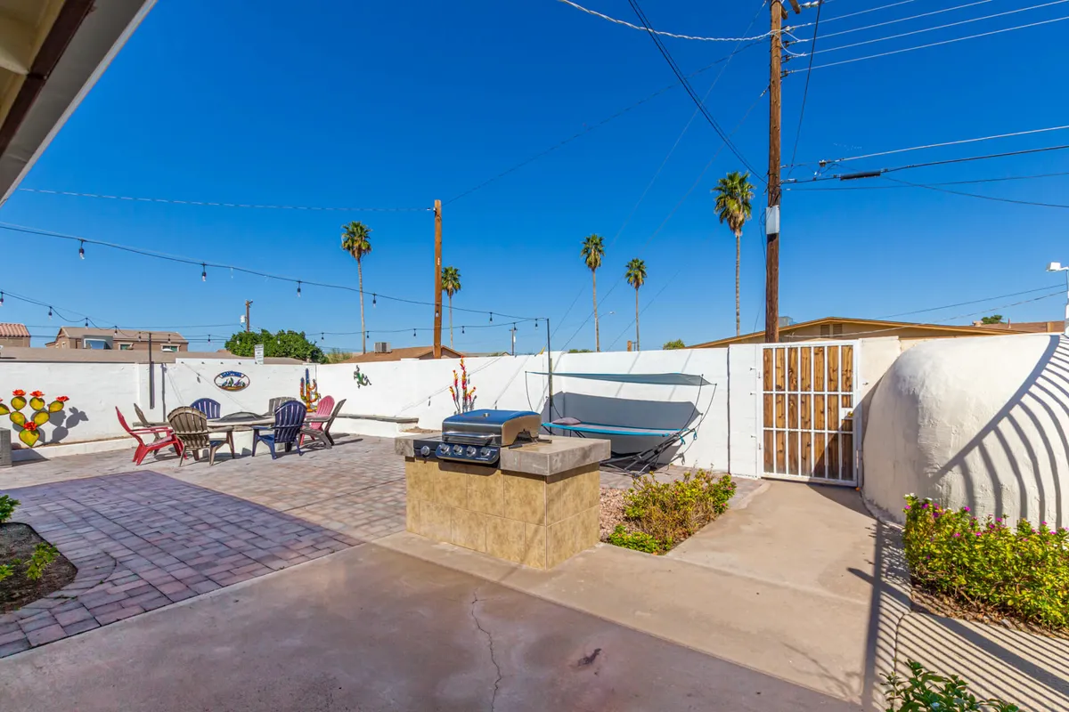 This exterior image captures a vibrant backyard featuring a grill, a comfortable sitting area, a hammock, palm trees, and decorative wall art. The scene is set for barbecuing, relaxing, and socializing, with string lights hinting at lively evening gatherings.