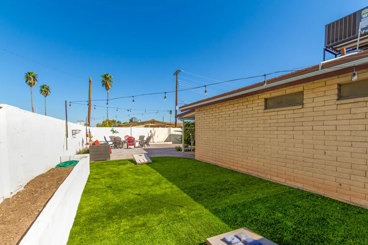 A vibrant backyard scene featuring a green lawn, an outdoor seating area, string lights, and palm trees. The setup suggests family gatherings, playing lawn games, and cheerful outdoor meals, with a mood that transitions well from daytime fun to evening relaxation.